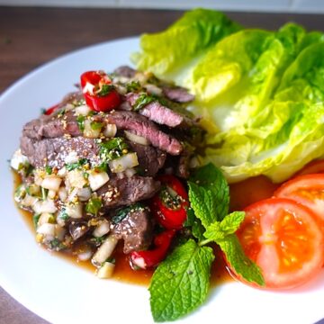 a white plate with grilled steak slices drizzled with thai spicy sauce garnished with lettuce & tomatoes