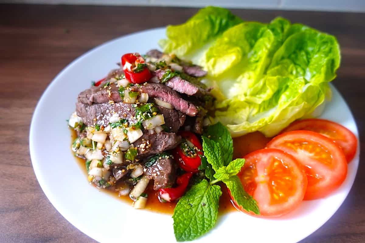 a white plate with grilled steak slices drizzled with thai spicy sauce garnished with lettuce & tomatoes