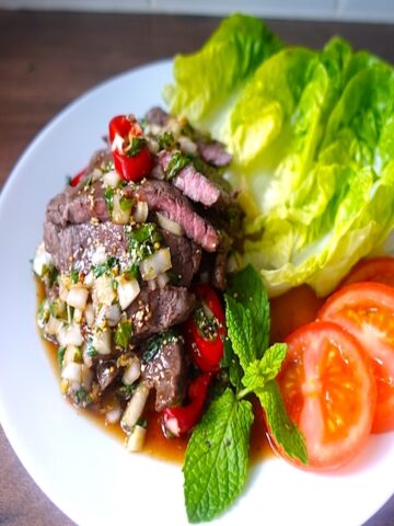 a white plate with grilled steak drizzled with thai spicy sauce served with lettuce & tomatoe