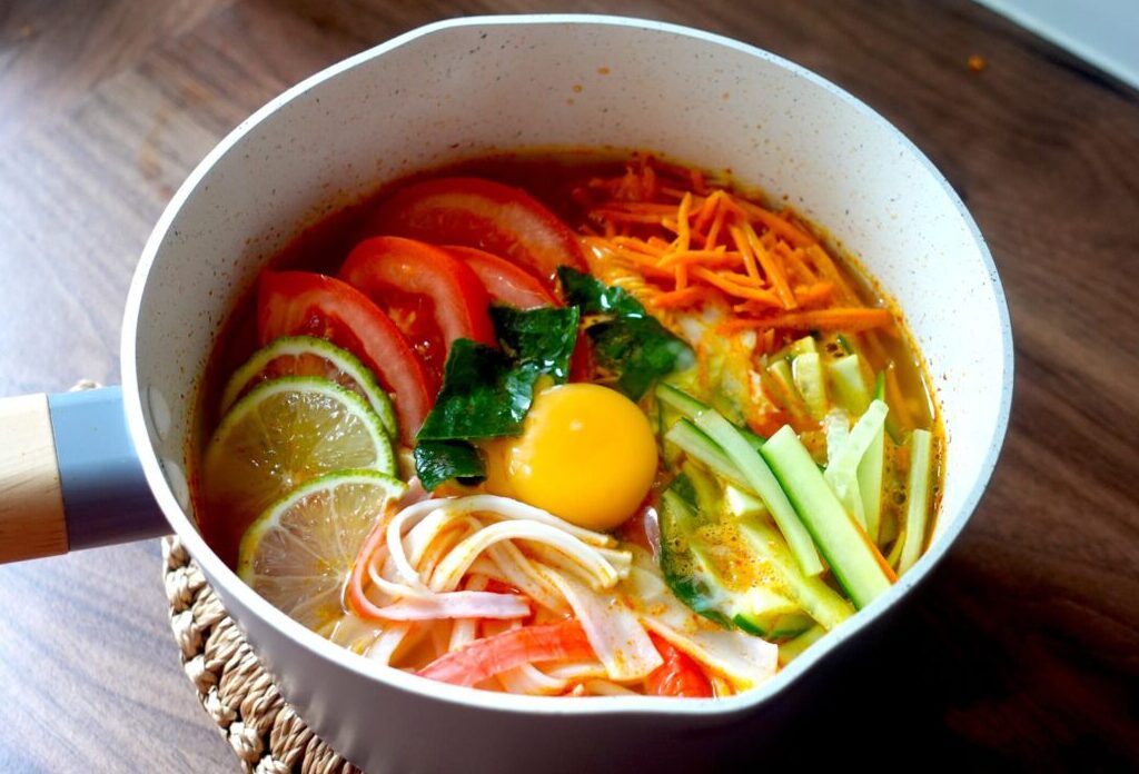 Tom Yum Noodles in a pot with tomato, cucumber, limes & surimi